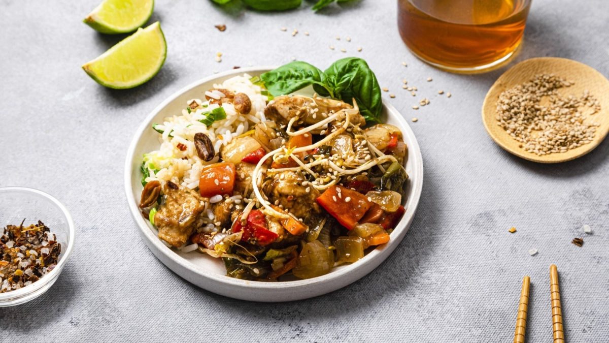 Veganes Chop Suey mit Reis serviert in einem weißen Teller. Rechts Stäbchen, Sesam und Öl. Links davon eine Limette und Chili-Salz. Der Hintergrund ist hellgrau.