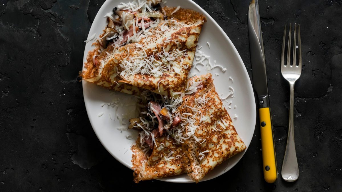 Draufsicht: Eine Teller mit zwei Crêpes mit Pilzen, Schinken und Käse auf dunklem Untergrund. Daneben liegen ein Messer und eine Gabel.