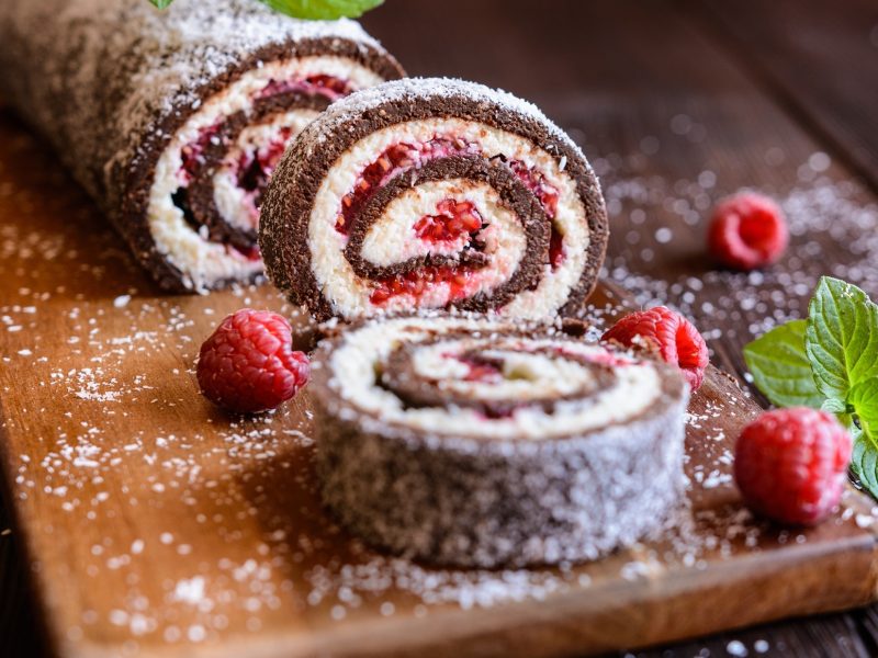 Frontal: Eine angeschnittene Biskuitrolle mit Schokolade und einer Fruchtmasse. Drum herum liegen frische Himbeeren.