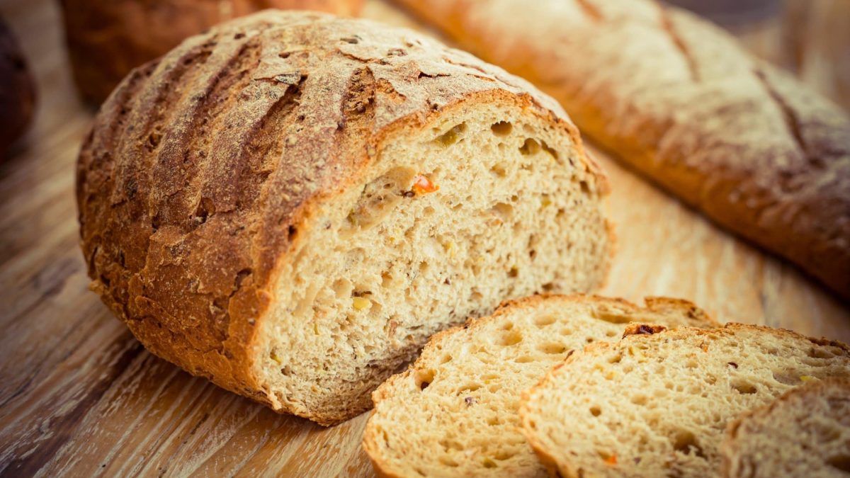 Draufsicht schräg: Ein angeschnittener Laib Ezekiel-Brot liegt auf einem Untergrund aus Holz. Davor liegen einige Scheiben des Brots aus der Bibel.