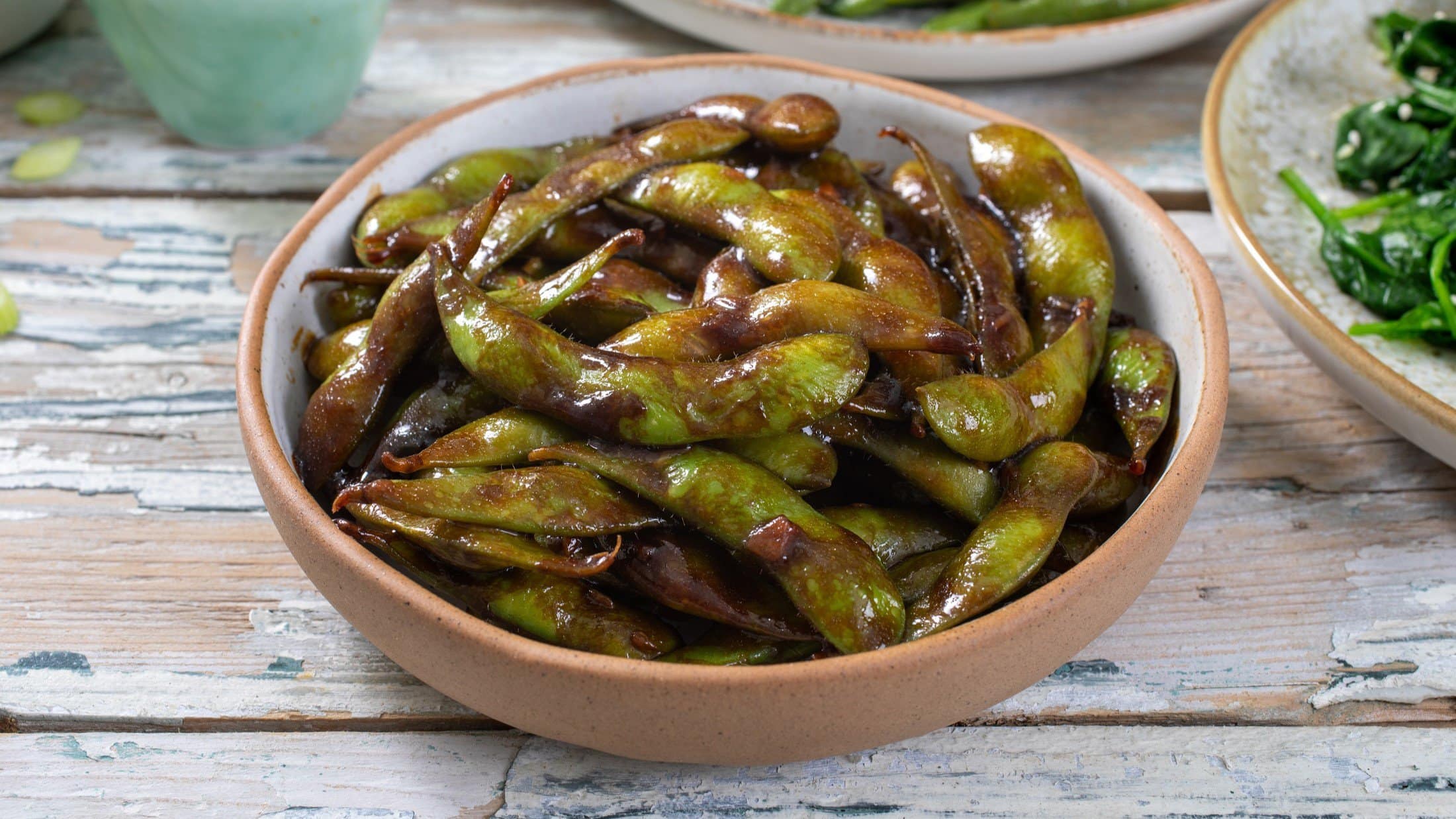 Draufsicht, schräg: Eine Snack-Schale gebratene Edamame mit Ingwer und Sojasauce auf einem hellen Untergrund aus Holz.