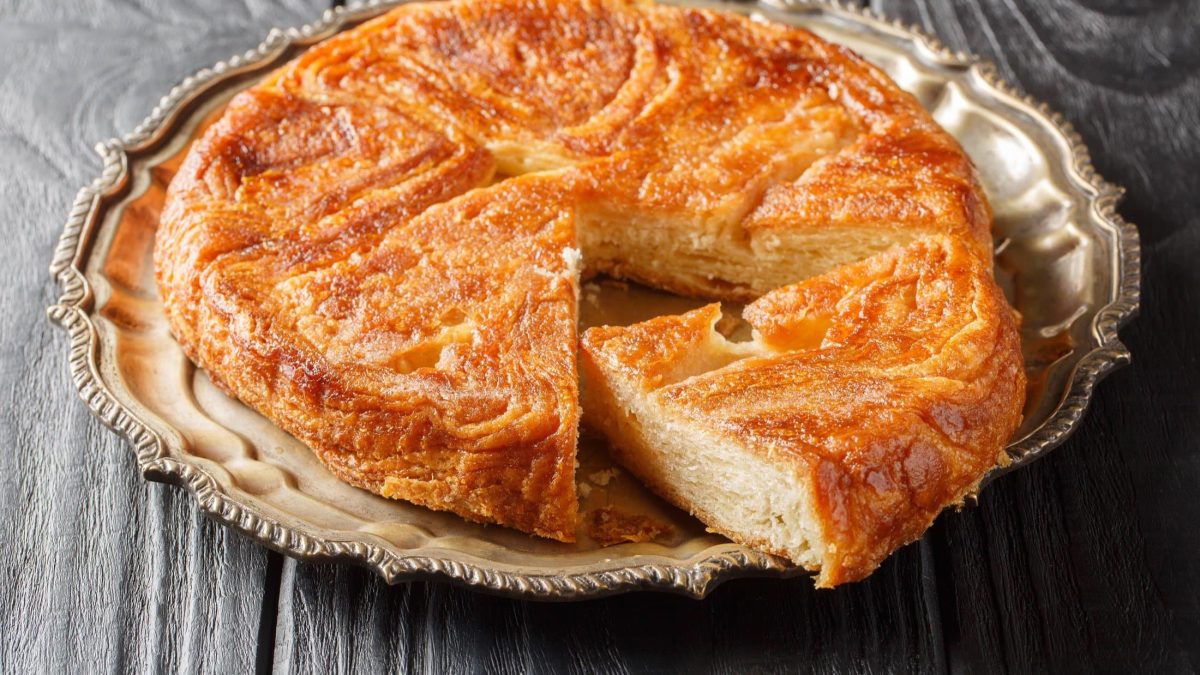 Draufsicht, schräg: Ein Kouign Amann auf einem Platte aus aus silber auf einem Holztisch. Es ist ein Bretonischer Butterkuchen.