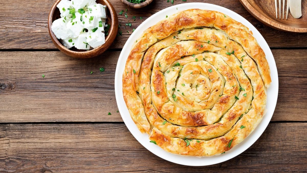 Die Feta-Spinat-Schnecke von oben auf einem dunklen Holztisch fotografiert. Daneben noch ein Schälchen mit Feta und eines mit gehackter Petersilie.