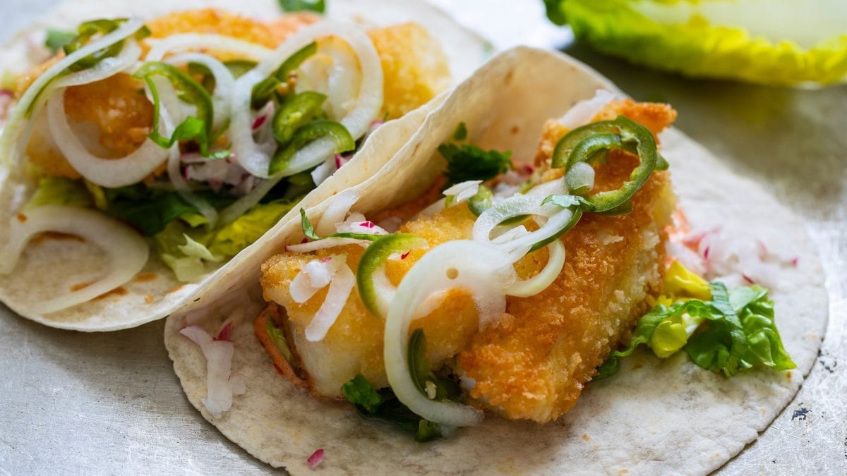 Zwei Fisch-Tacos auf einem hellgrauen Tisch nah fotografiert.