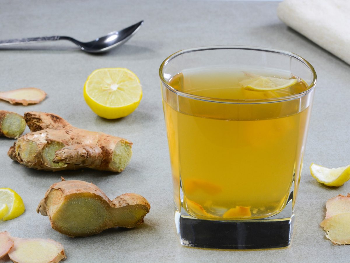 Ein Glas mit dem Ingwerwasser und Ingwer selbst auf einer grauen Tischplatte von der Seite fotografiert.