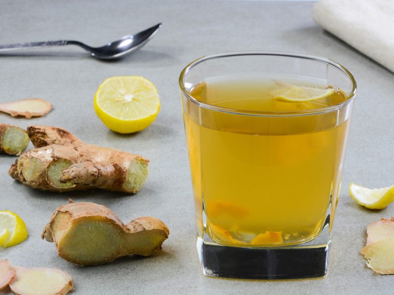 Ein Glas mit dem Ingwerwasser und Ingwer selbst auf einer grauen Tischplatte von der Seite fotografiert.