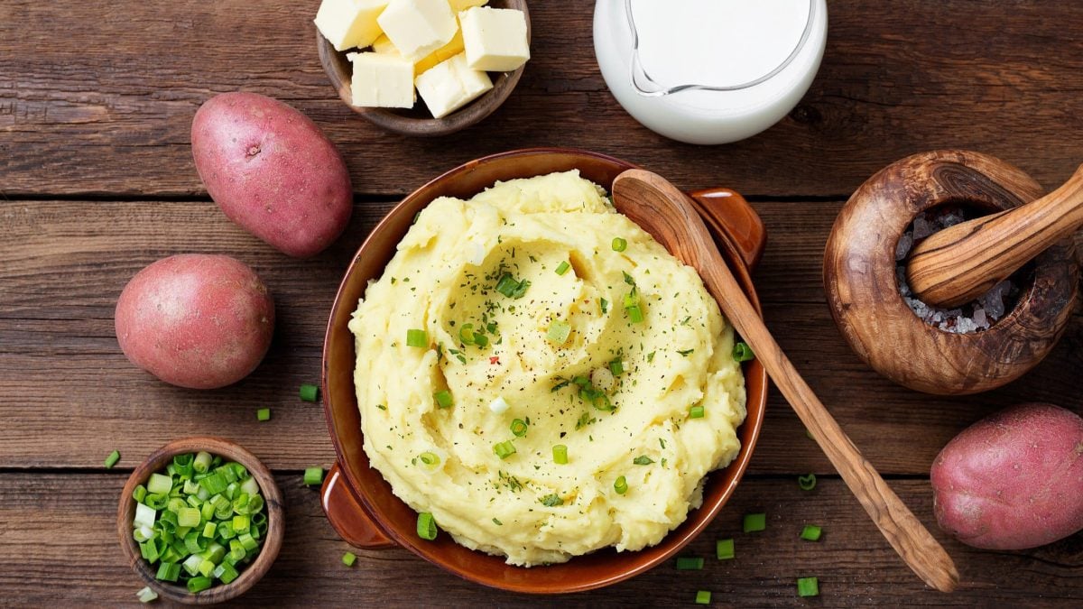 Draufsicht: Untergrund aus Holz. Eine runde Tonschlüssel Kartoffelpüree mit Rettich. Drum herum liegen rotschalige Kartoffeln, Schnittlauchröllchen, Butterwürfel und ein Holzlöffel.