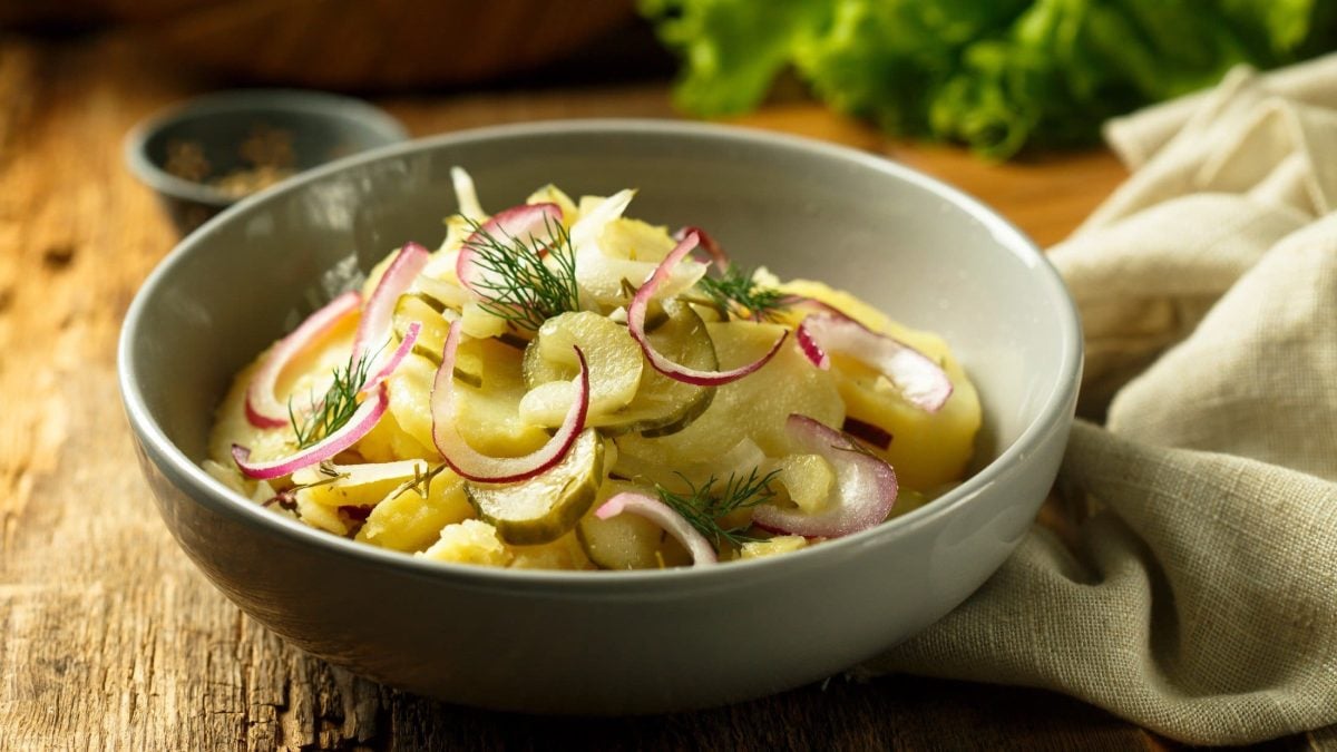 Draufsicht, schräg: Eine runde, graue Schüssel Kartoffelsalat mit Gewürzgurken steht auf einem Holztisch. Drum herum liegen ein Tuch und Gemüse.