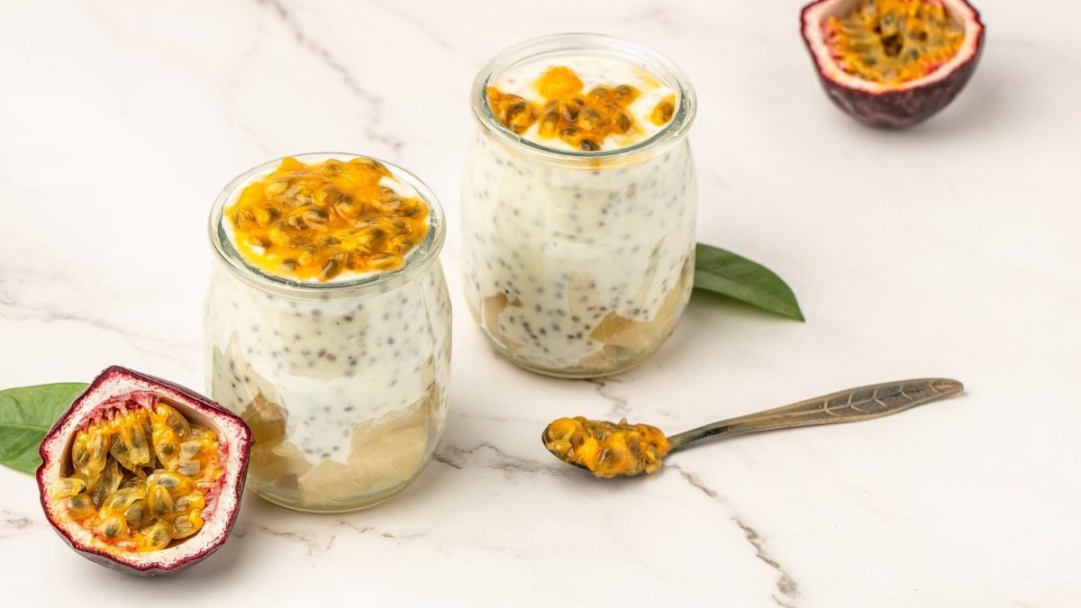 Zwei Gläser mit dem Kokos-Chia-Pudding auf einem hellen Marmortisch. Daneben zwei halbierte Passionsfrüchte und zwei grüne Blätter. Alles von schräg oben fotografiert.