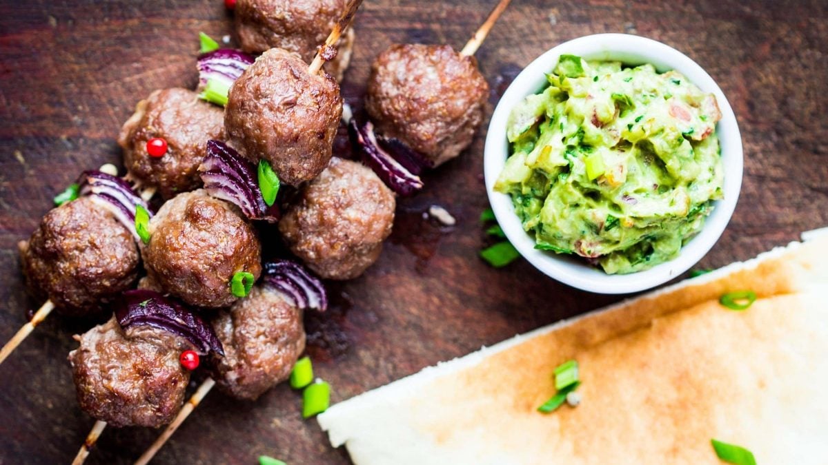Draufsicht: Drei Spieße Lammbuletten liegen auf einem Untergrund aus Holz. Daneben ist eine Schüssel mit einem Avocado-Dip.