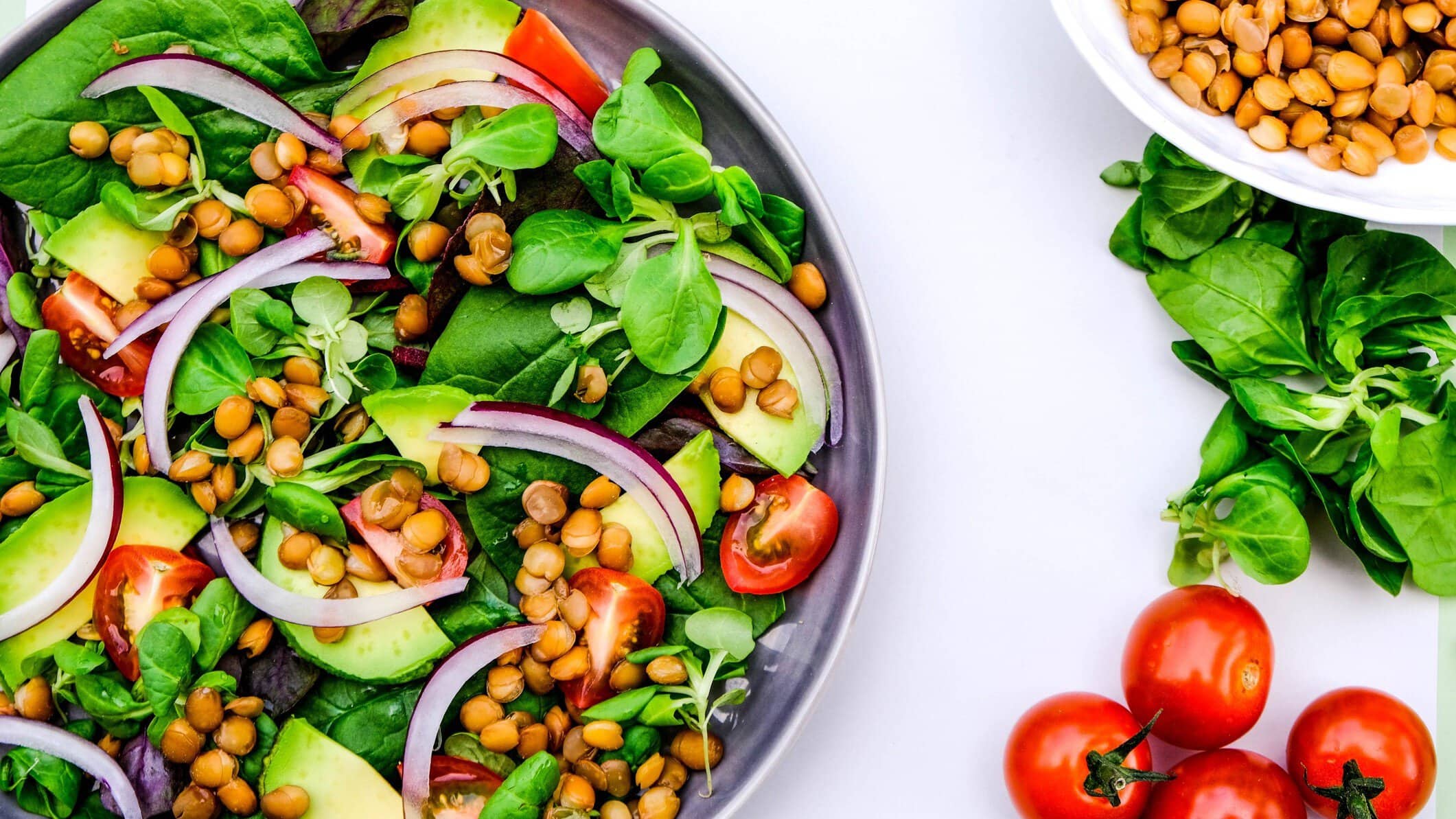 Draufsicht: Ein halbe Schüssel Linsen-Avocado-Salat. Daneben liegen kleine Tomaten, Spinatblätter und Linsen.