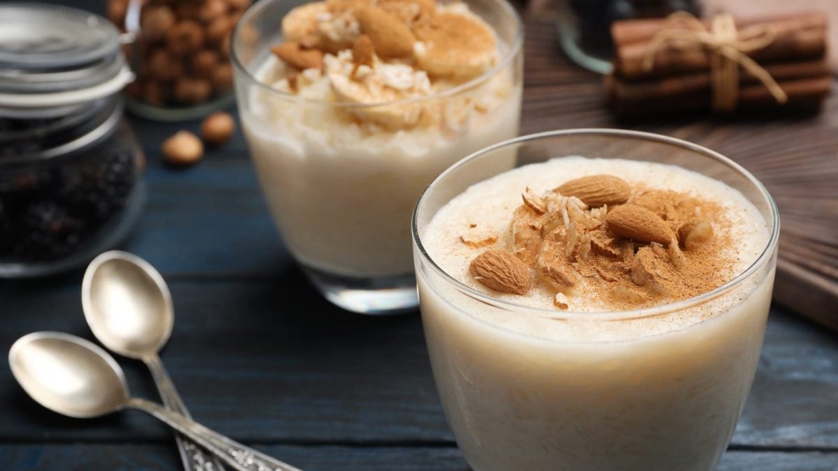 Zwei Gläser mit dem Mandel-Milchreis auf einem dunkelblauen Holztisch mit zwei silbernen Löffeln sowie Gewürzen.