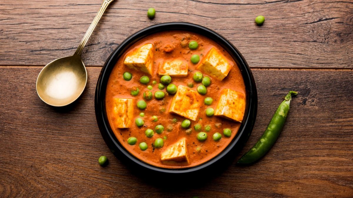 Matar Paneer in einer schwarzen Schüssel auf braunem Hintergrund. Links davon ein Löffel und Erbsen auf dem Hintergrund verteilt.
