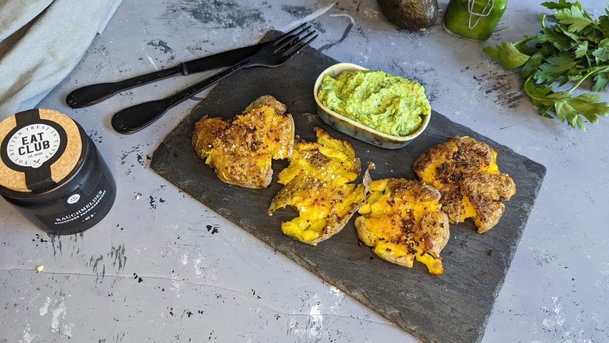 Eine Schieferplatte mit den Kartoffeln und dem Dip auf einem grauen Tisch mit Avocado und Petersilie. Alles von schräg oben fotografiert.