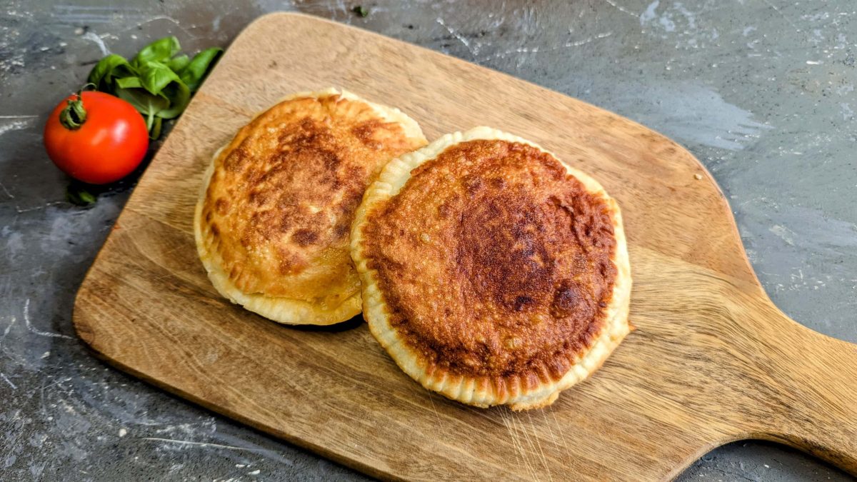 Zwei frittierte Pizzen auf einem Holzbrett und grauem Untergrund. Daneben Tomate und Basilikum. Alles von schräg oben fotografiert.