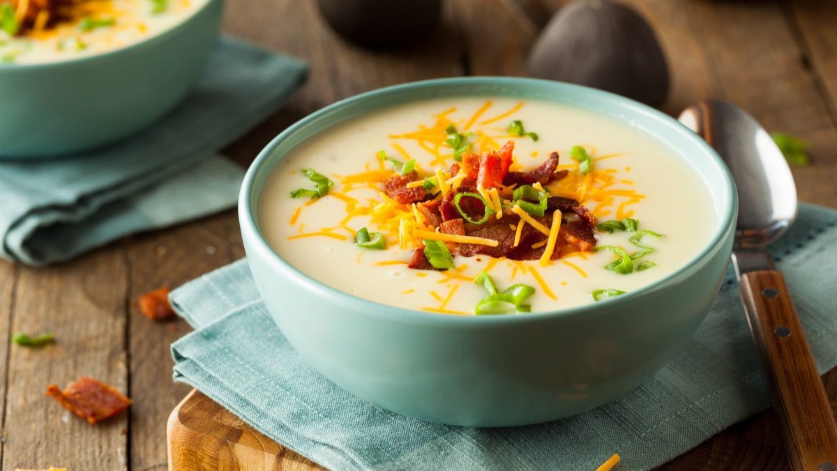 Draufsicht, schräg: Eine Raclette-Suppe mit Speck in einer hellblauen Schüssel. Sie steht auf einem Tuch aus Leinen. Daneben liegt ein Suppenlöffel.