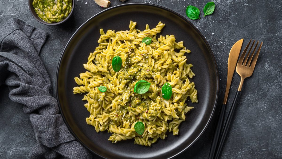 Ein schwarzer Teller mit dem Pasta-Gericht und schwarz-goldenem Besteck sowie grauem Tuch und Schale mit dem Pesto. Alles auf dunklem Untergrund von oben fotografiert.