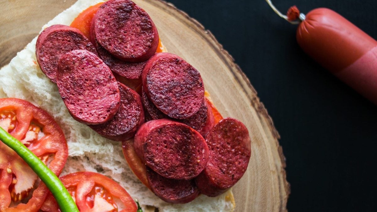 Draufsicht: eine aufgeklappte Sucuk-Stulle auf einem Holzbrett. Neben der Sucuk liegen auf dem Weißbrot eine Tomate und eine grüne Peperoni.