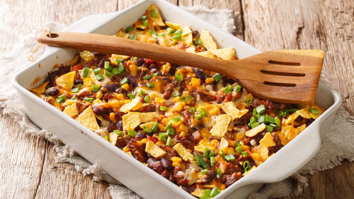 Eine weiße Auflaufform mit dem Taco-Auflauf auf einem hellen Holztisch von schräg oben fotografiert.