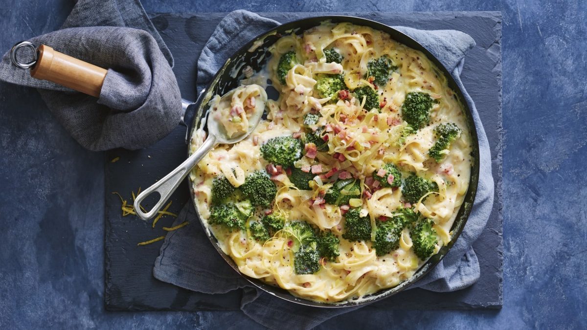 Tagaliatelle mit Käsesauce und Brokkoli in einer schwarzen Pfanne auf einem dunkelgrauen Geschirrtuch auf einer grauen Schieferplatte. Der Hintergrund ist ebenfalls dunkelgrau.