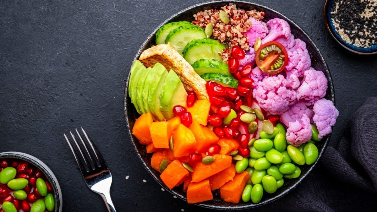 Vegane Regenbogen-Bowl auf schwarzem Untergrund, daneben Zutaten und eine Gabel.