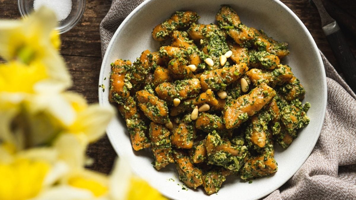 Draufsicht: Vegane Süßkartoffel-Gnocchi mit Spinat-Pesto und Pinienkernen auf weißem Teller mit Holzuntergrund und unscharfen gelben Pflanzen daneben.