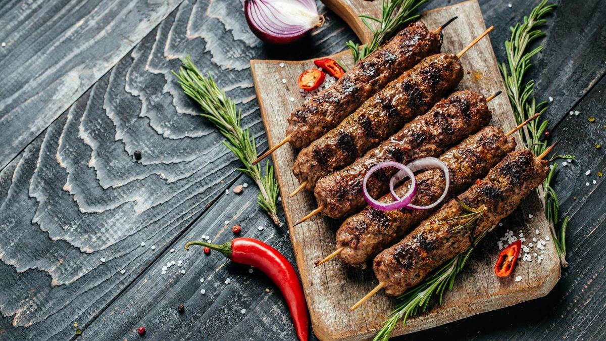 Mehrere Walnussköfte-Spieße auf einem Holzbrett von oben auf einem dunklen Tisch fotografiert. Daneben Rosmarinzweige, Chili und rote Zwiebel.