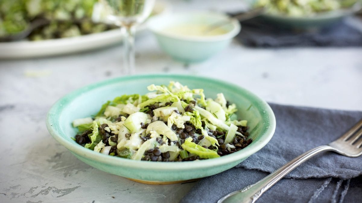 Draufsicht schräg: ein Teller mit einem Linsensalat mit Apfel, der auf einem Tisch steht. Im Hintergrund ist ein Glas.
