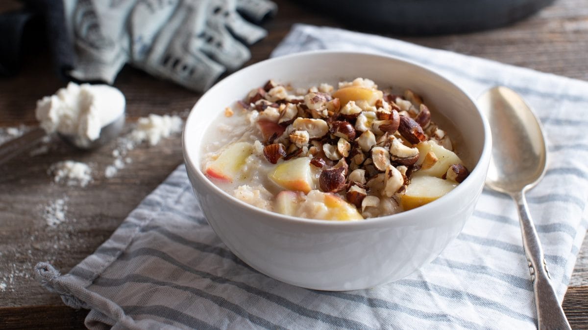 Draufsicht, schräg: Ein Apfel-Porridge mit gerösteten Nüssen. in einer Schüssel liegt auf einem Tuch, dass auf einem Holztisch liegt. Daneben sind ein Löffel, eine Hantel und ein Handschuh sowie ein weißes Pulver zu sehen.
