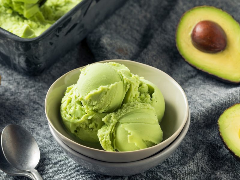 Drei Kugeln cremiges Avocado-Eis in einer grauen Schale, daneben frische Avocadohälften auf einer Stoffunterlage mit zwei Löffeln.