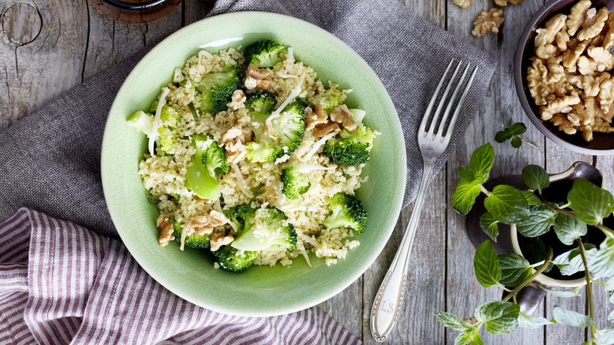 Bulgur mit Brokkoli in einer hellgrünen Schüssel auf einem grauen Geschirrtuch auf hölzernem Hintergrund. Rechts ein Schüsselchen mit Walnüssen und frische Minze.