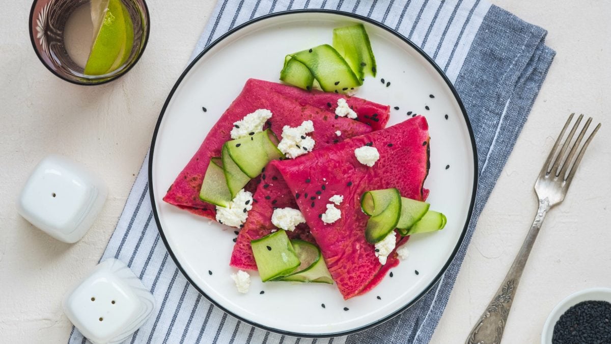 Rote Bete Crêpes mit Sesam, Hüttenkäse und Gurke auf einem weißen Teller auf einem blau-weiß-gestreiftem Geschirrtuch. Rechts davon eine Gabel, links auf dem Bild Salz- und Pfefferstreuer. Links oben ein Glas Wasser,