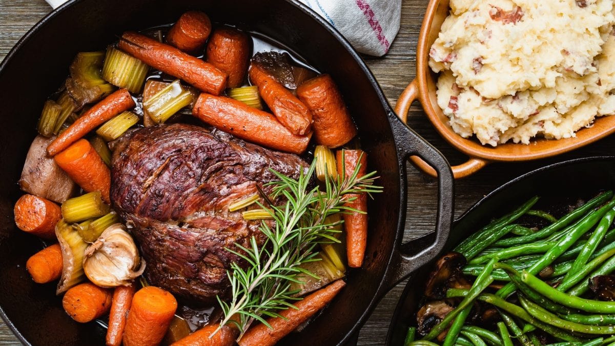 Draufsicht: Ein schwarzer gusseiserner Topf, in dem ein Braten und Schmorgemüse in einer Flüssigkeit ist, steht auf einem Untergrund aus Holz. Es handelt sich um einen italienischen Brasato al Barolo.