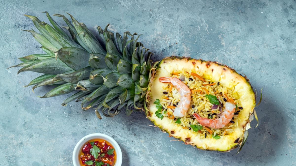 Eine ausgehöhlte Ananas mit dem Reisgericht auf einem hellblauen Tisch. Alles direkt von oben fotografiert.