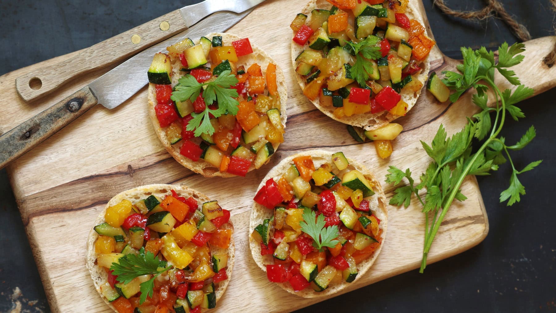Ratatouille-Brötchen auf einem hölzernen Schneidebrett. Links Besteck. Der Hintergrund ist dunkelgrau.