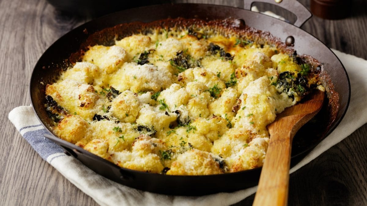 Draufsicht, schräg: Ein Blumenkohl-Grünkohl-Gratin in einer gusseisernen Pfanne. Die steht auf einem Tuch, dass auf einem Untergrund aus Holz ist. In der Pfanne ist ein großer Löffel aus Holz.