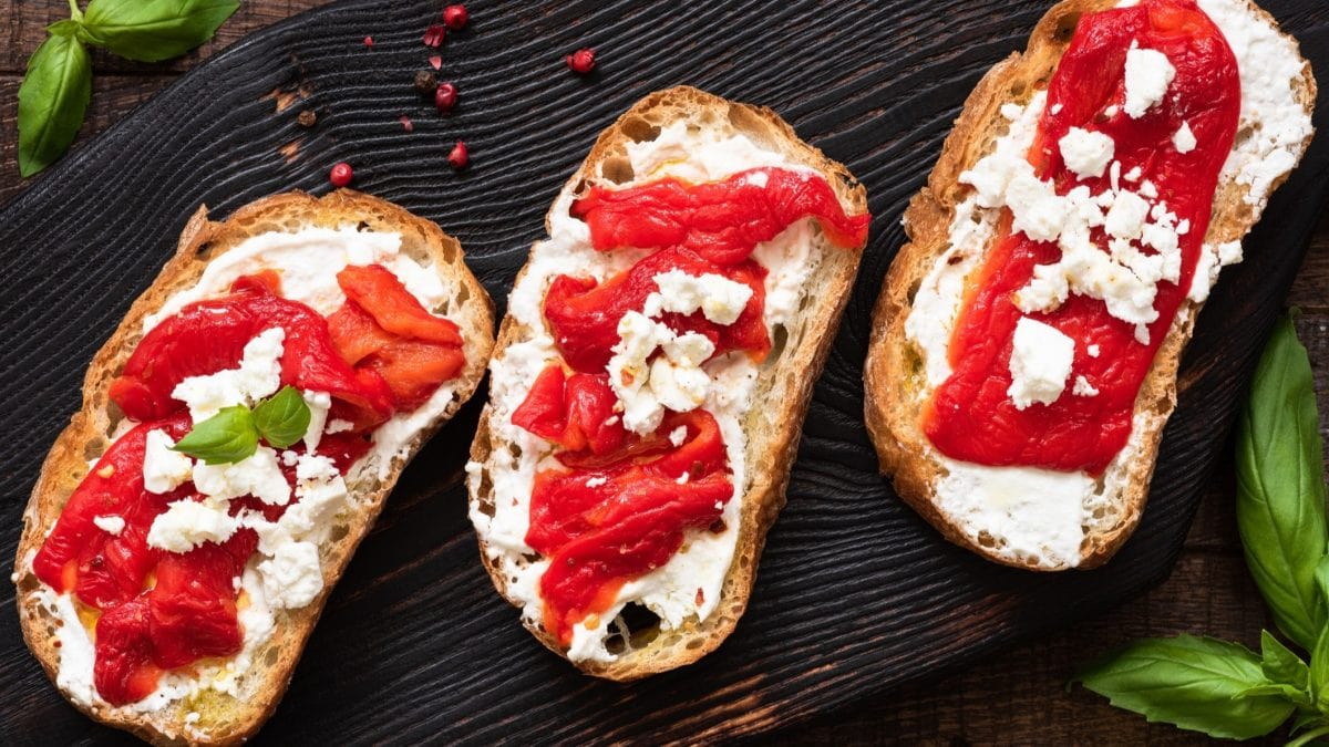 Draufsicht: drei Bruschetta mit gerösteter Paprika und Ricotta auf schwarzem Untergrund. Drum herum liegen frische Basilikumblätter.