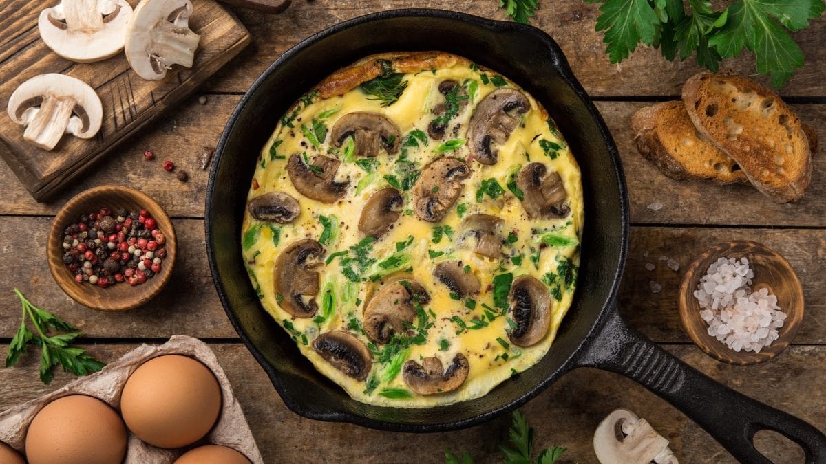 Draufsicht: Eine dunkle Pfanne in der eine Champignon-Frittata ist. Die steht auf einem rustikalem Tisch aus Holz. Drum herum liegen viele Zutaten für das Rezept.