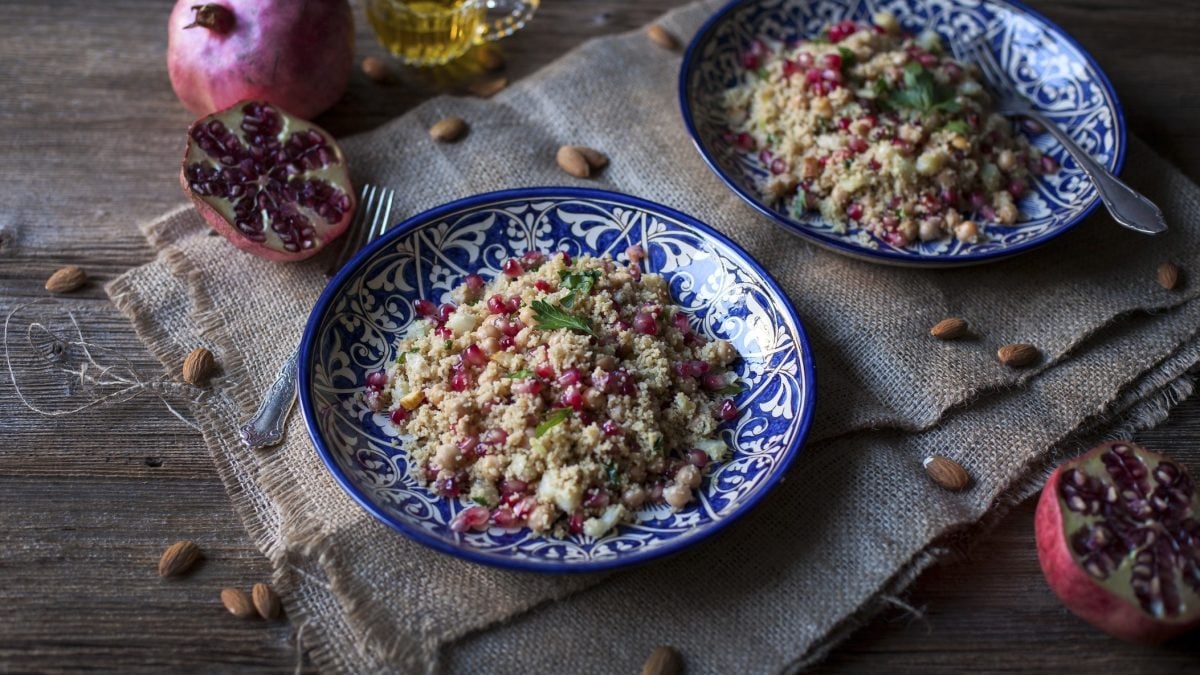 Draufsicht: Zwei Teller Couscous-Salat mit Granatapfel auf einem Untergrund aus Holz. Drum herum liegen Mandeln und ein aufgeschnittener Granatapfel.