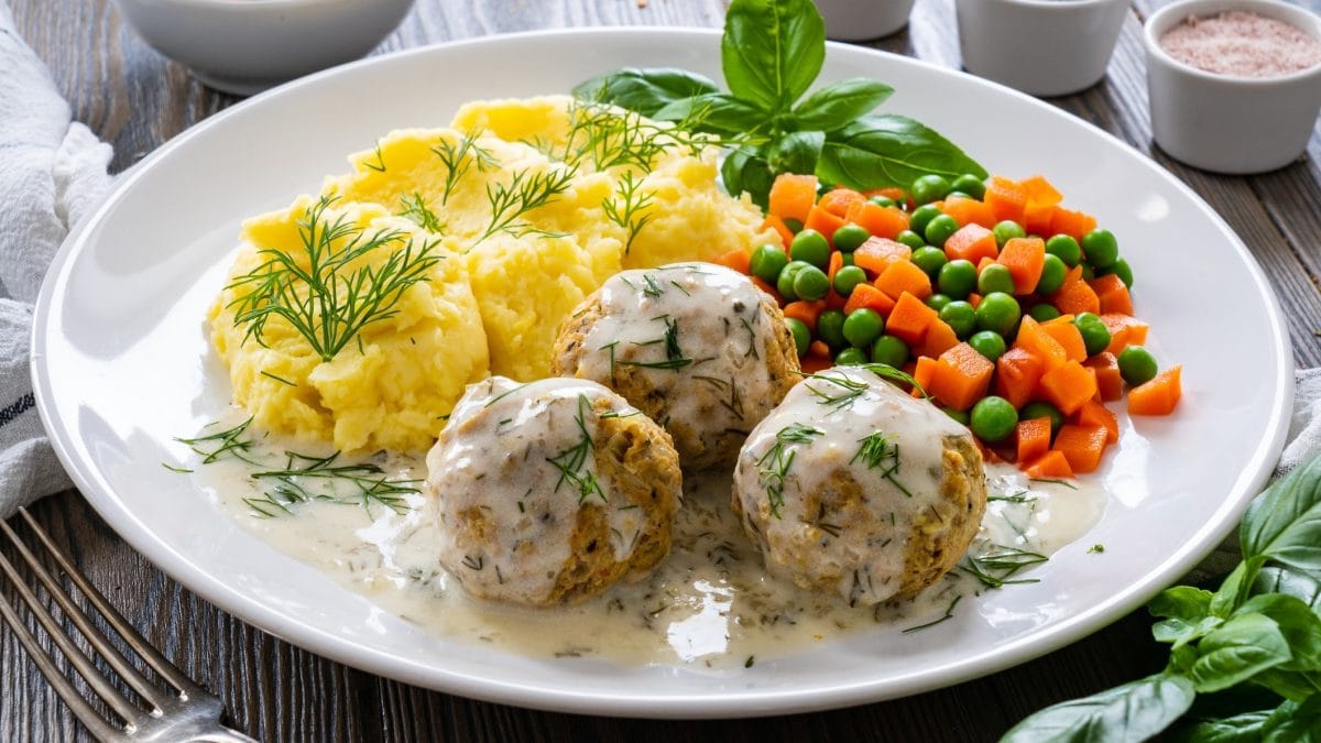 Draufsicht, schräg: Ein weißer Teller mit Fleischbällchen in Dillsauce sowie Kartoffelpüree und Möhren-Erbsen-Gemüse steht auf einem Holztisch.