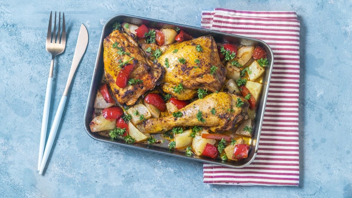 Draufsicht: Auf einem hellblauen Untergrund steht einen ofenfeste Auflaufform mit Hähnchen mit Paprika-Gemüse. Daneben liegen ein rot-weiß-gestreiftes Handtuch und Besteck.