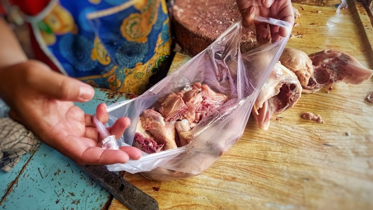 Draufsicht: Ein Mensch mit einer Schürze steht auf einem asiatischen Markt. In der Hand hält er eine offene Einkaufstüte. In der sind Karkassen vom Geflügel.