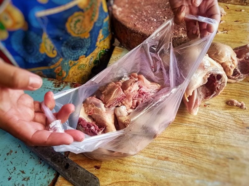 Draufsicht: Ein Mensch mit einer Schürze steht auf einem asiatischen Markt. In der Hand hält er eine offene Einkaufstüte. In der sind Karkassen vom Geflügel.