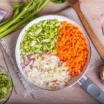 Draufsicht: Auf einer Arbeitsplatte in einer Küche liegt eine Pfanne mit gleichmäßig geschnittenem Gemüse. Es handelt sich um ein Mirepoix. Drum herum liegen Küchenhandtücher und klein geschnittenes Gemüse.