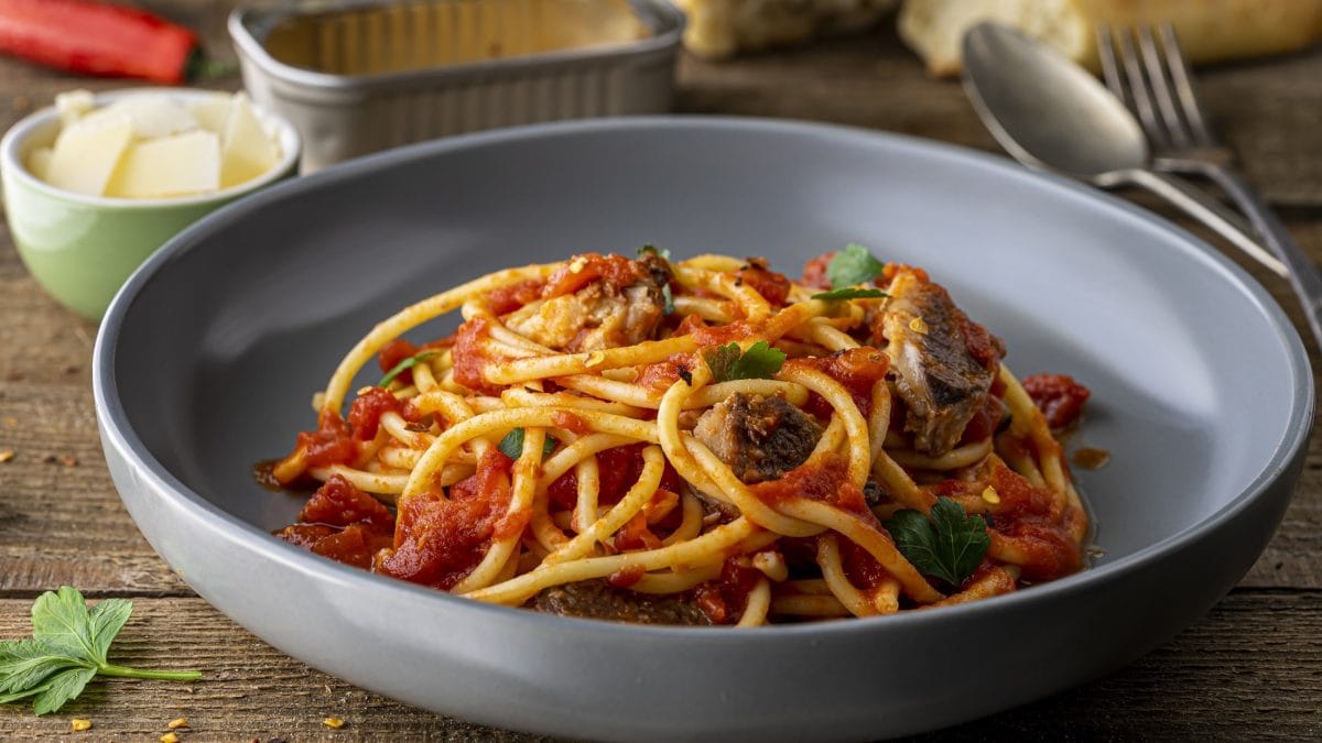 Frontal: Ein Teller Pasta mit Sardinien. auf einem Holztisch. Im Hintergrund sind Butter, Weißbrot und einen Dose, in der die Sardinen waren.