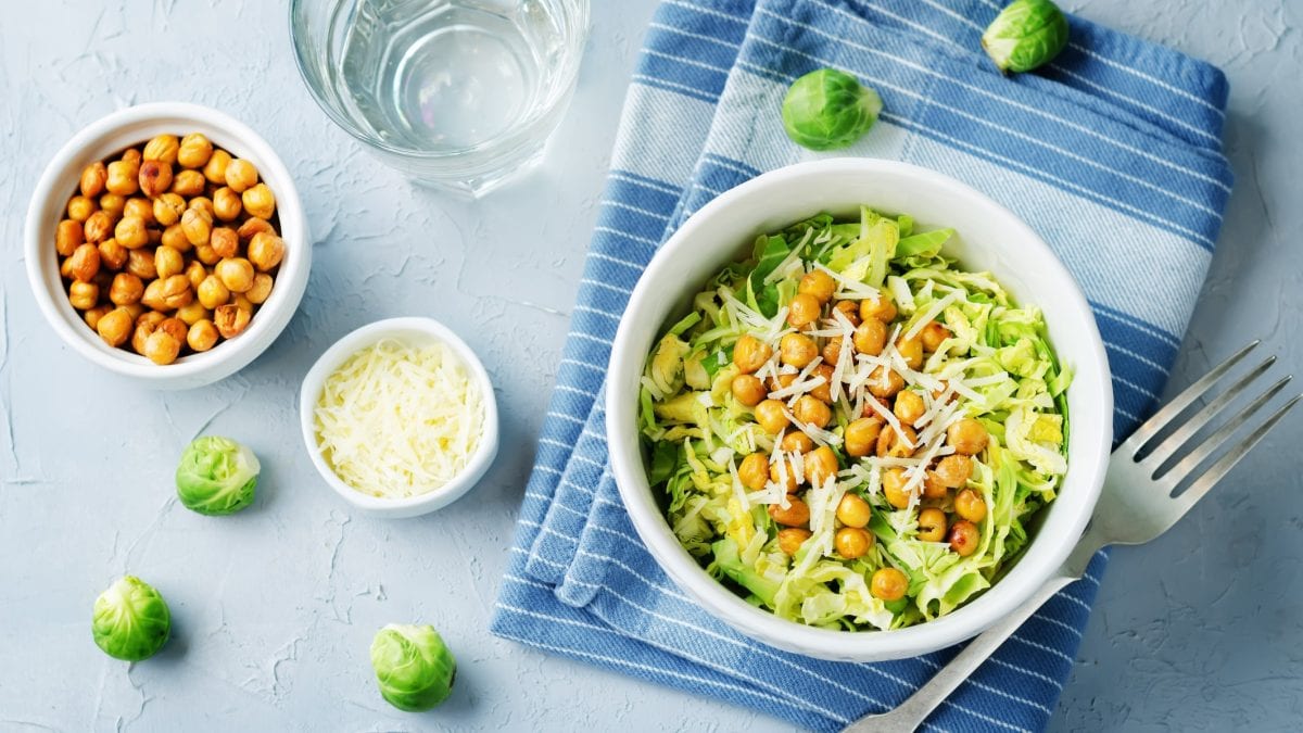 Draufsicht: auf hellblauem Untergrund ligt ein hellblaues Tuche auf dem eine Schale mit Rosenkohl-Kichererbsen-Salat steht. Drum herum sind eine Schale mit Kichererbsen, eine Schale mit geriebenem Käse sowie roher Rosenkohl.