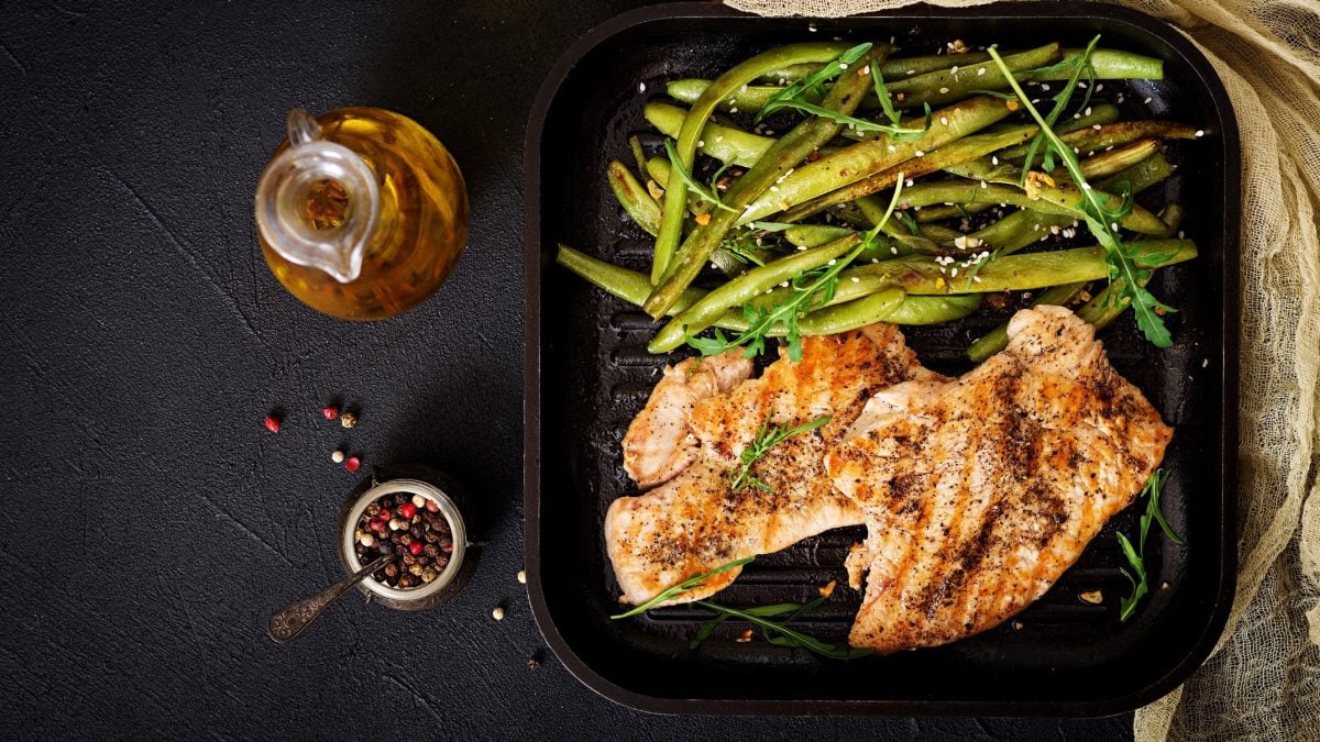 Draufsicht: schwarzer Untergrund, auf dem eine quadratische Grillpfanne mit Sesam-Hähnchen mit Bohnen. Drum herum liegen Gewürze.