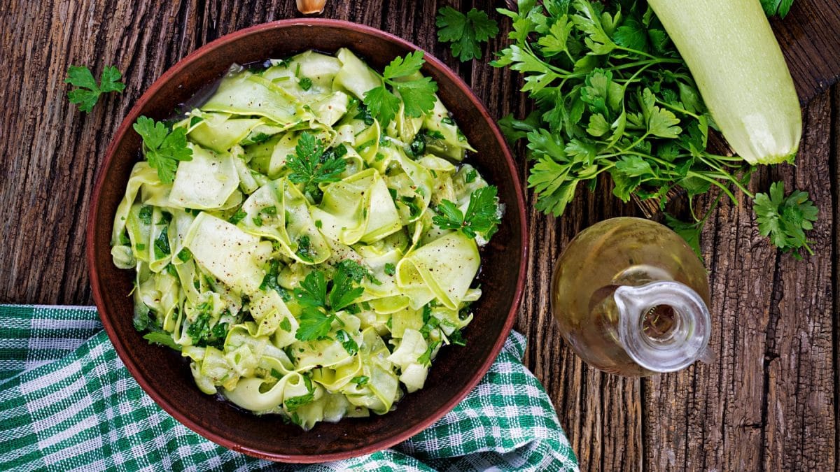 Eingelegter Zucchini-Salat in einer Schüssel mit Petersilie darauf und daneben auf Holzuntergrund. Daneben Ölflasche, Küchentuch, Petersilie und eine helle Zucchini.