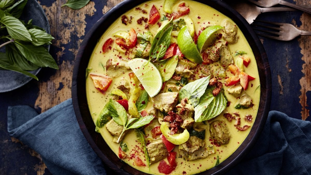 Entencurry in einer dunkelbraunen Schüssel, links davon Basilikum, rechts auf dem Bild Besteck. Der Hintergrund ist hölzern. Unten auf dem Bild ein dunkelgraues Geschirrtuch.