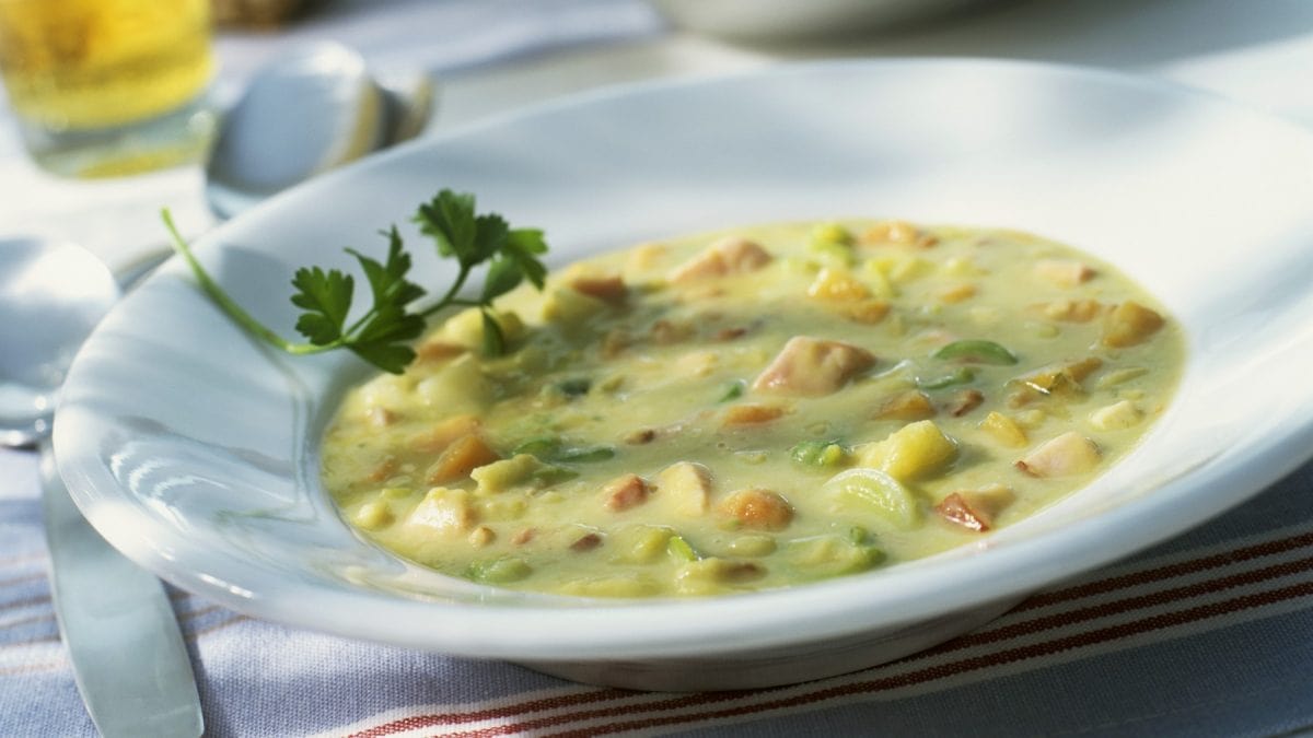 Erbsensuppe mit Würstchen in einem weißen Suppenteller auf einem Tisch. Links davon ein Löffel.